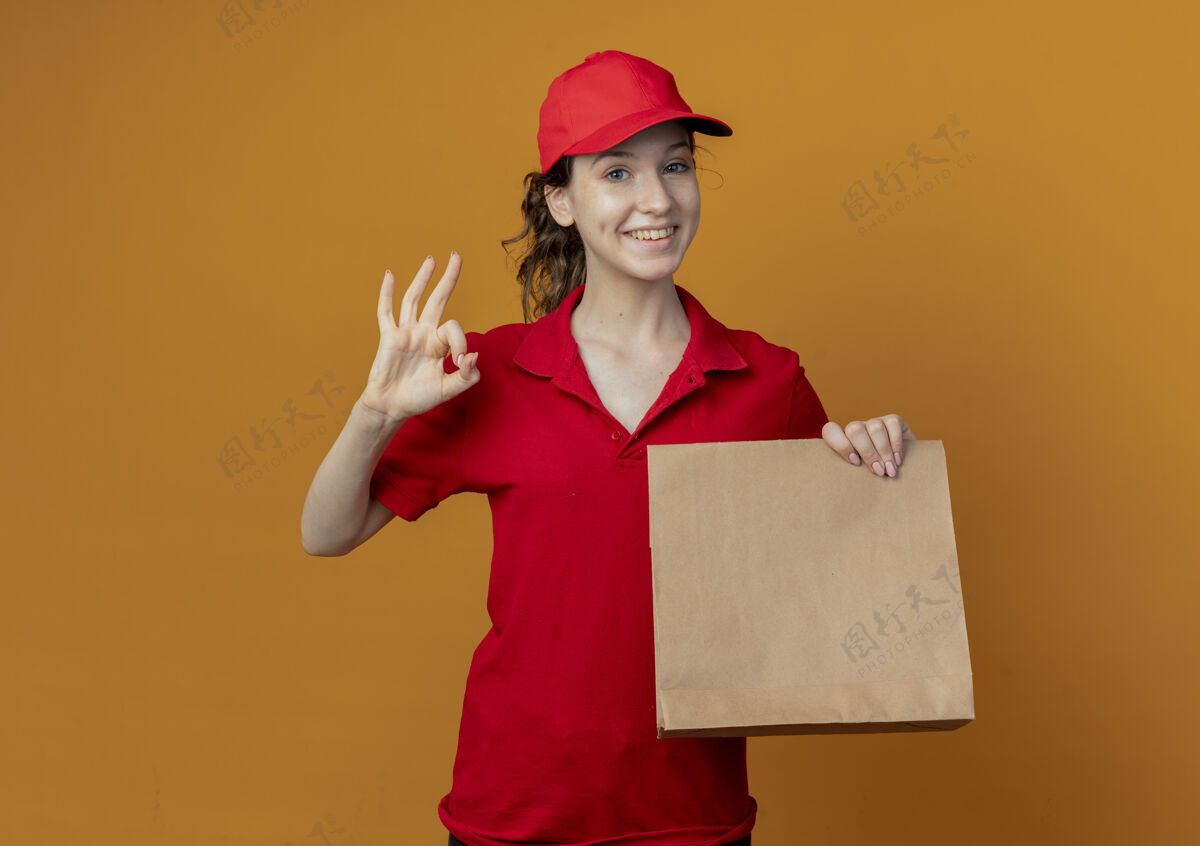 包装微笑着的年轻漂亮的送货女孩 穿着红色制服 戴着帽子 展示着纸包装和doingok标志 隔离在橙色背景上 留有复印空间制服交货好