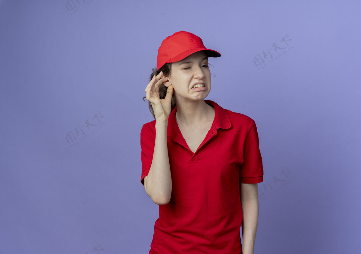 漂亮恼怒的年轻漂亮的送货女孩穿着红色制服 戴着帽子 看着侧面 把手放在耳朵上 紫色背景上 留着复印空间恼火制服帽子
