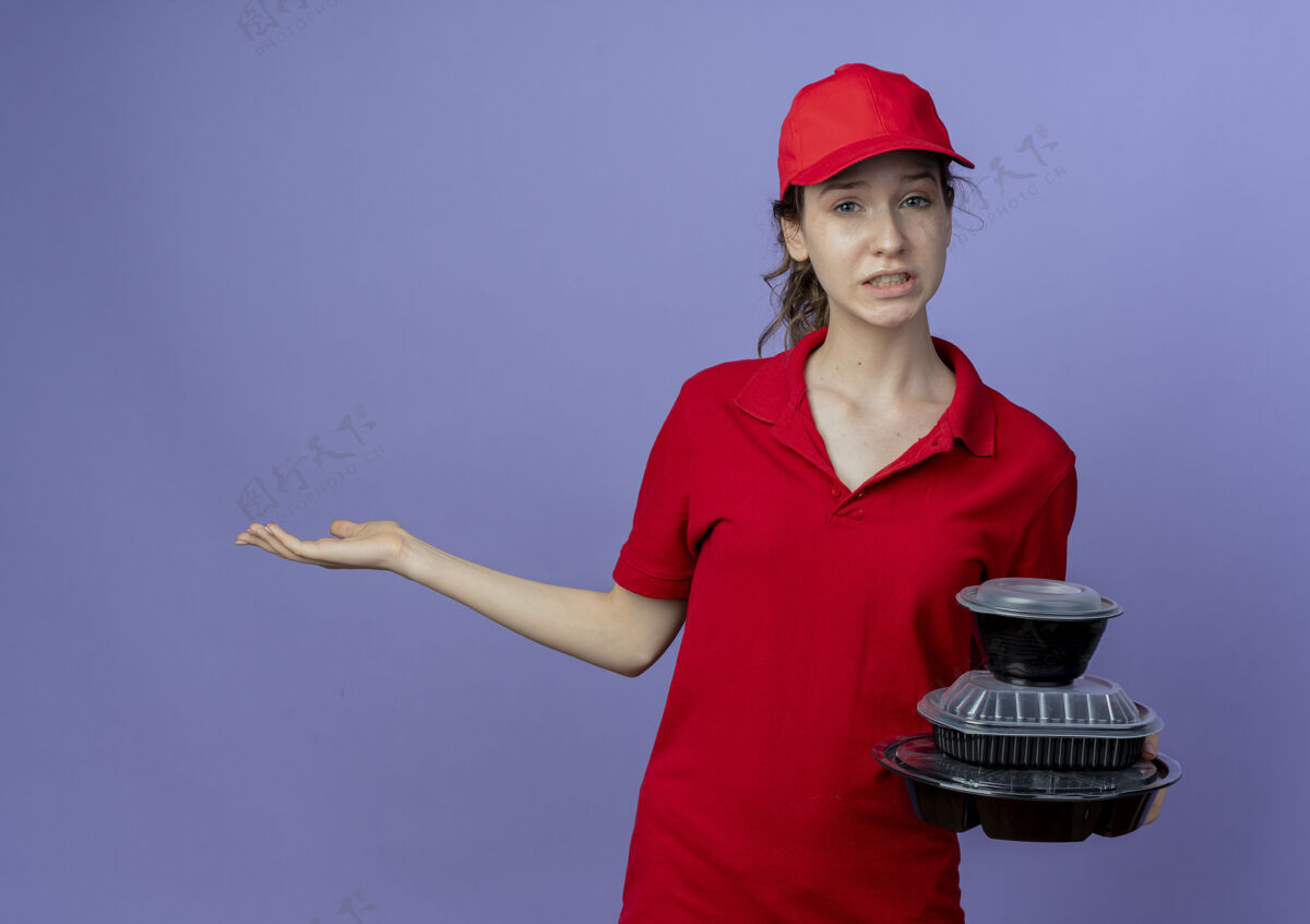 年轻年轻漂亮的送货女孩 穿着红色制服 戴着帽子 手里拿着食物容器 手上空空的 背景是紫色的 还有复印空间容器帽子空