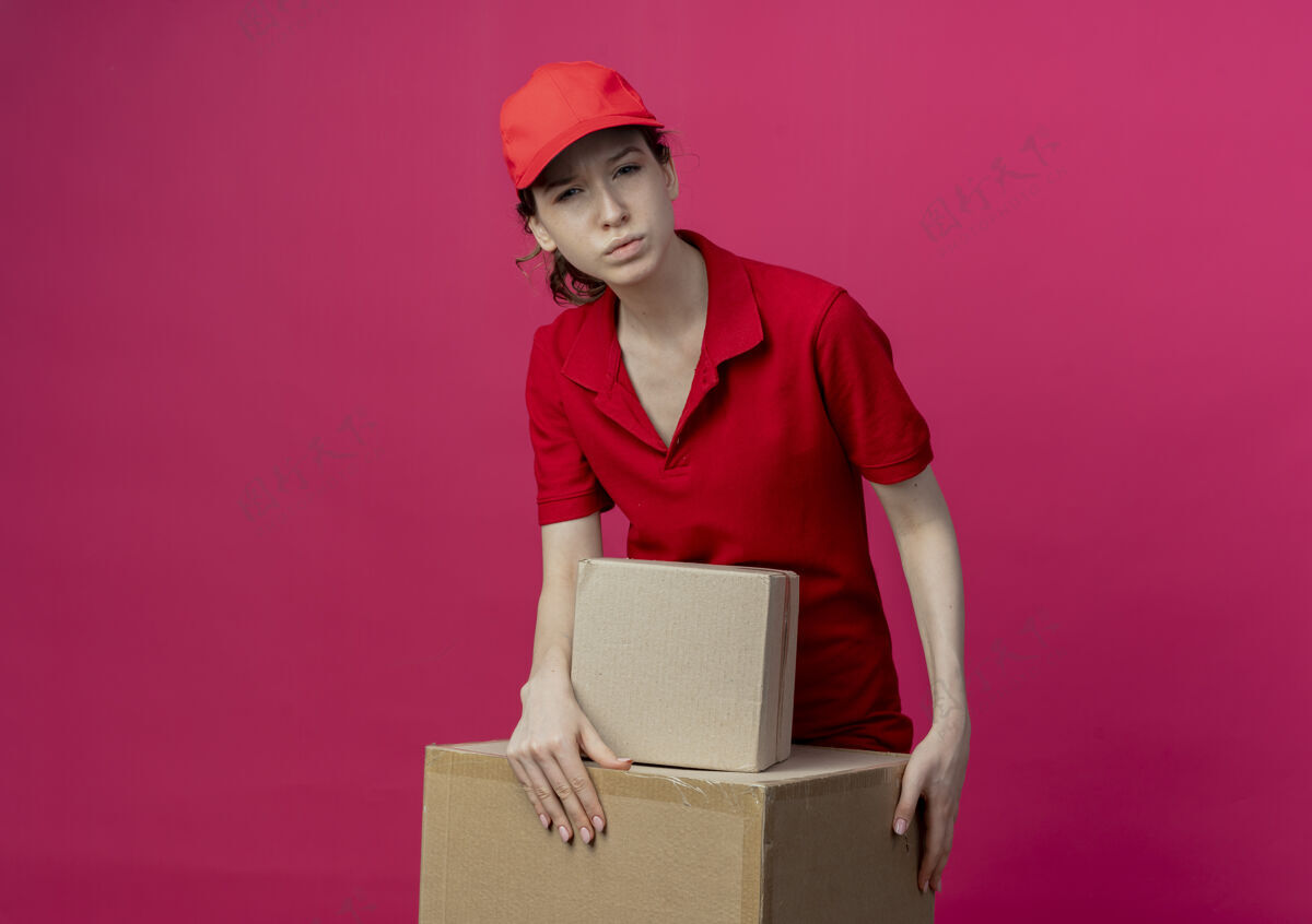 手体贴的年轻漂亮的送货女孩 穿着红色制服 戴着帽子 手放在深红色背景的纸箱上 有复印空间制服盒子深红色