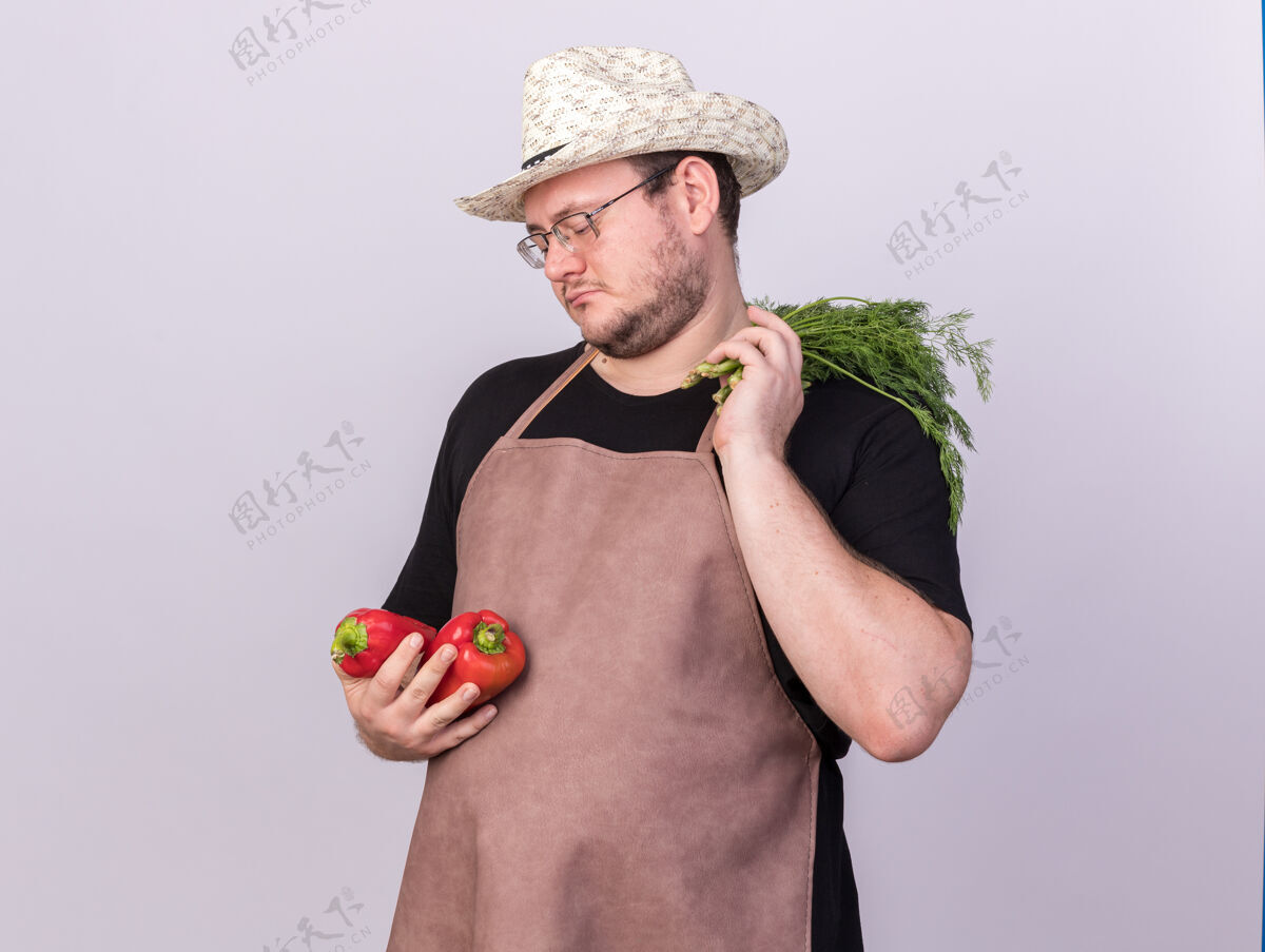 人悲伤的年轻男园丁戴着园艺帽拿着辣椒和莳萝隔离在白墙上胡椒持有立场