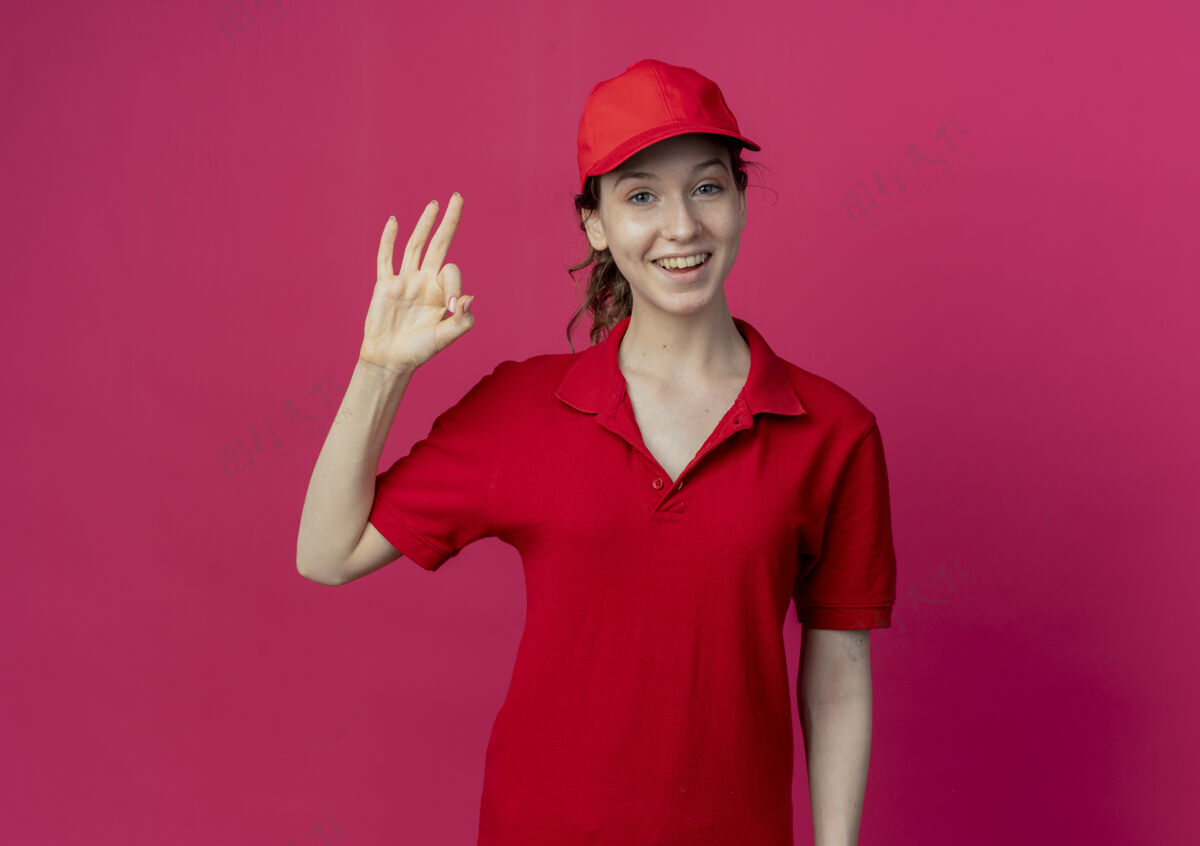 年轻快乐的年轻漂亮的送货女孩 穿着红色制服 戴着帽子 在深红色的背景上做着“ok”的标志 并留有复印空间欢乐交货红色