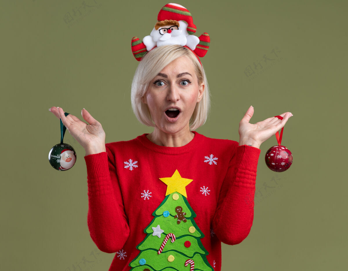 圣诞饰品令人印象深刻的中年金发女人戴着圣诞老人的头带和圣诞毛衣拿着圣诞饰品看着橄榄绿背景上孤立的相机圣诞快乐印象金发