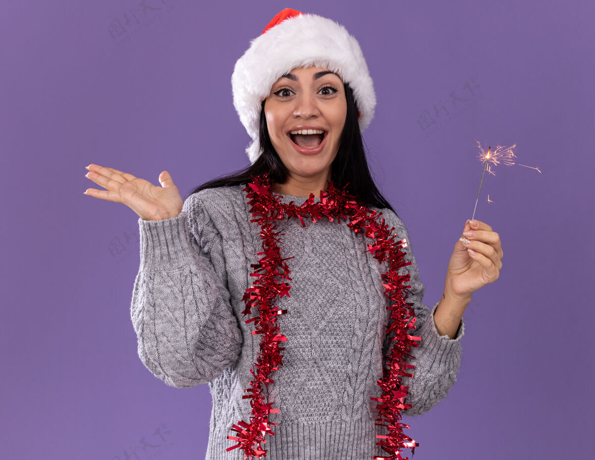年轻令人印象深刻的年轻白人女孩戴着圣诞帽 脖子上戴着金箔花环 手里拿着节日火花灯 看着镜头 在紫色背景上孤立着一只空手花环印象圣诞节