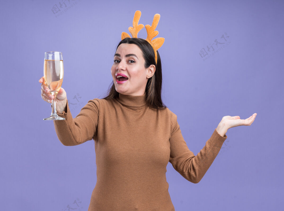 女孩兴奋的年轻漂亮女孩穿着棕色毛衣 戴着圣诞发箍 手拿香槟 手孤立在蓝色背景上香槟毛衣摊开