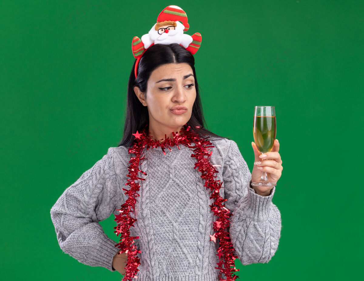 香槟可疑的年轻白人女孩戴着圣诞老人的头带 脖子上戴着金属丝的花环 手放在腰上 手拿着 看着绿色背景上孤立的一杯香槟周围花环女孩