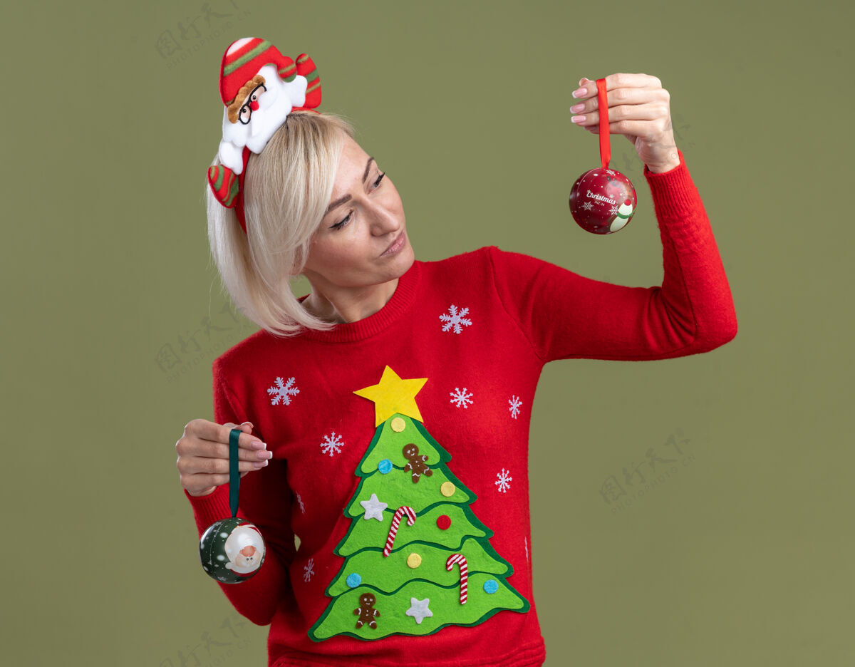 圣诞快乐可疑的中年金发女人戴着圣诞老人的头带和圣诞毛衣拿着圣诞饰品看着一个孤立的橄榄绿背景一件女人中年