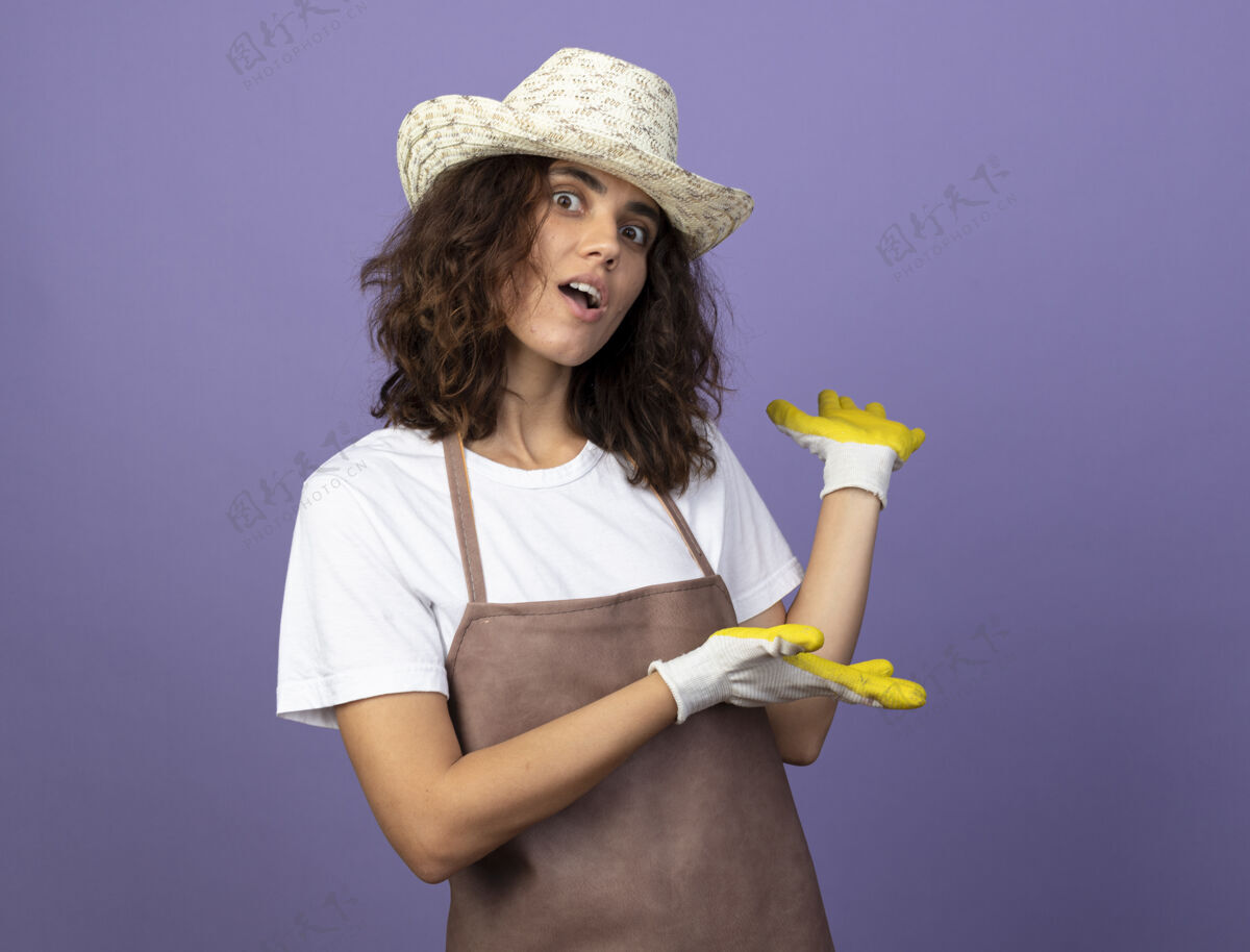 人惊讶的年轻女园丁穿着制服戴着园艺帽和手套点在后面穿花园女
