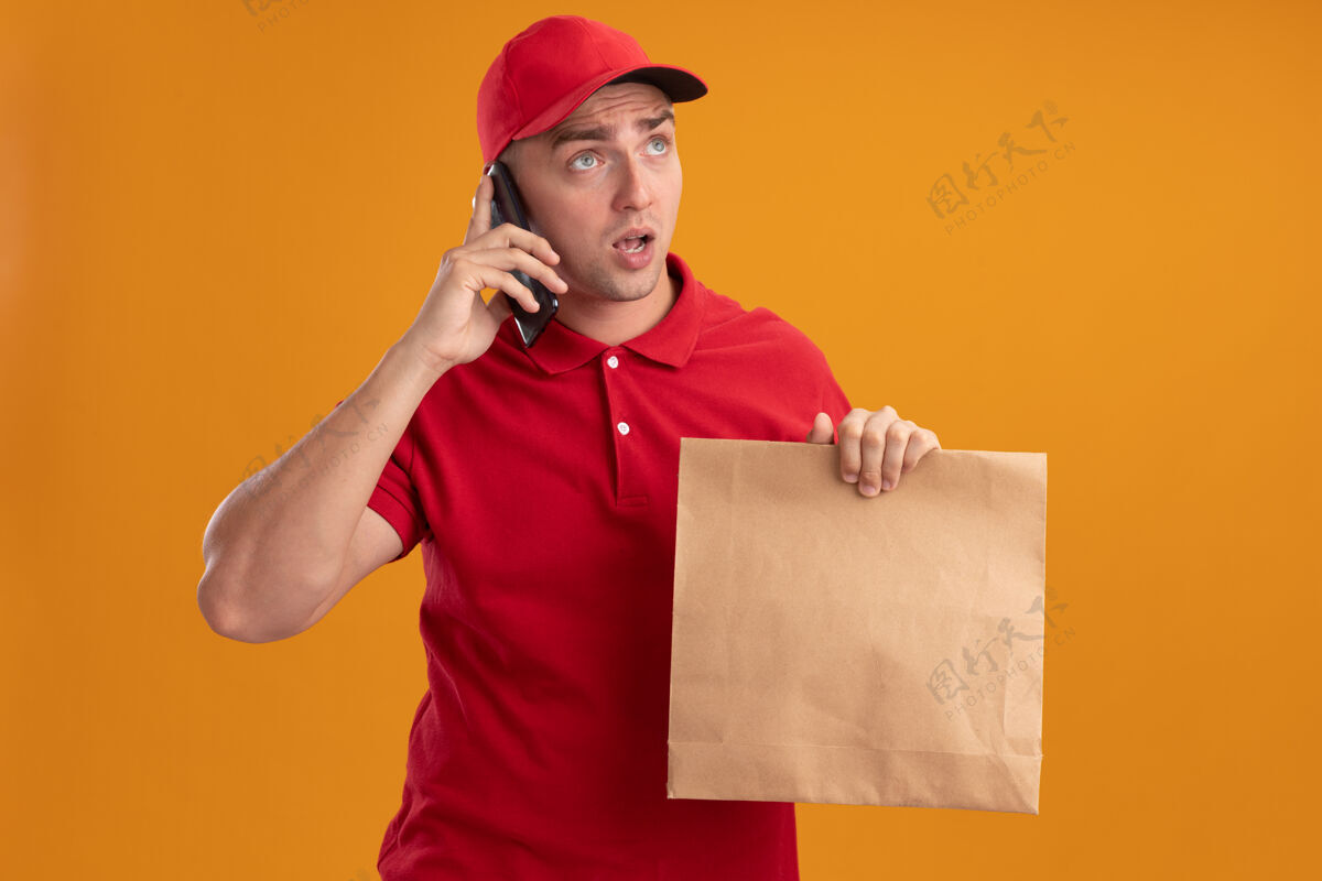 电话惊讶地看着身边穿着制服戴着帽子拿着纸食品包的年轻送货员讲电话隔离在橙色的墙上人包装说话