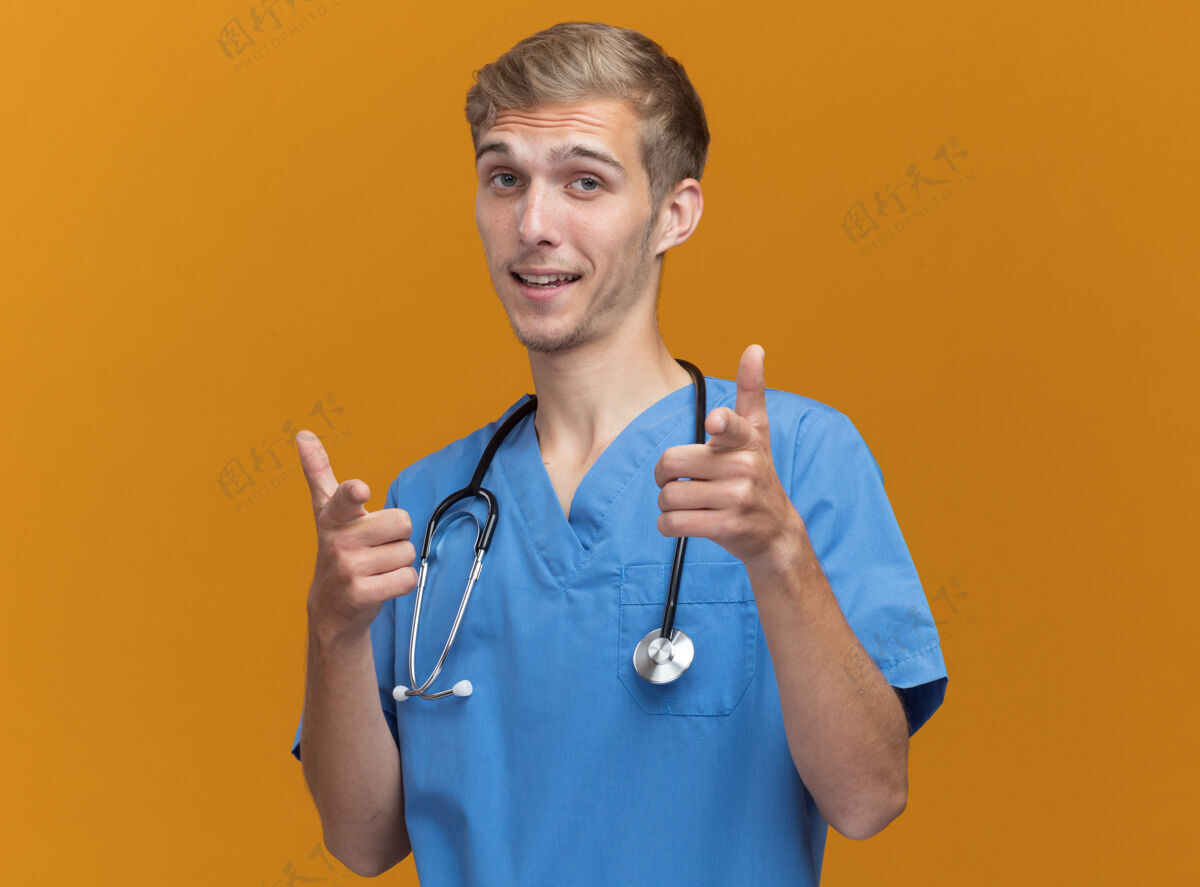 人微笑着的年轻男医生穿着医生制服 手持听诊器指着隔离在橙色墙上的摄像机点脸年轻人