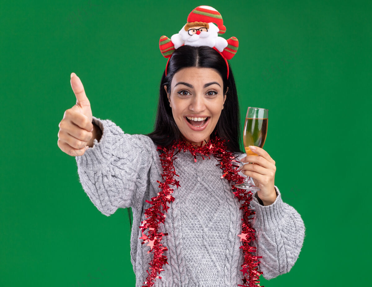 香槟快乐的年轻白人女孩戴着圣诞老人的头带 脖子上戴着金箔花环 手里拿着一杯香槟 看着相机 在绿色的背景上孤立地竖起大拇指金属片圣诞老人圣诞老人