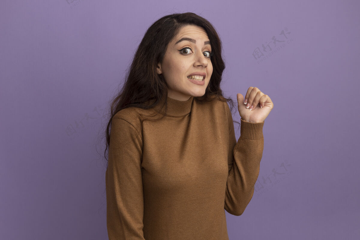 点关心的年轻漂亮女孩穿着棕色高领毛衣点在后面 隔离在紫色的墙上和复制空间衣服关心脸