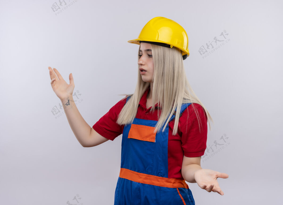 金发困惑的年轻金发工程师建设者女孩穿着制服 张开双臂 在孤立的空白处看着左边女孩年轻手臂