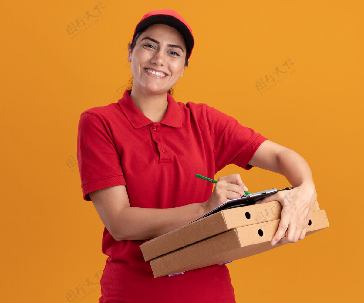 脸微笑着的年轻送货女孩看着前面穿着制服 戴着帽子 在剪贴板上写着什么 就在隔离在橙色墙上的比萨饼盒上感觉微笑披萨