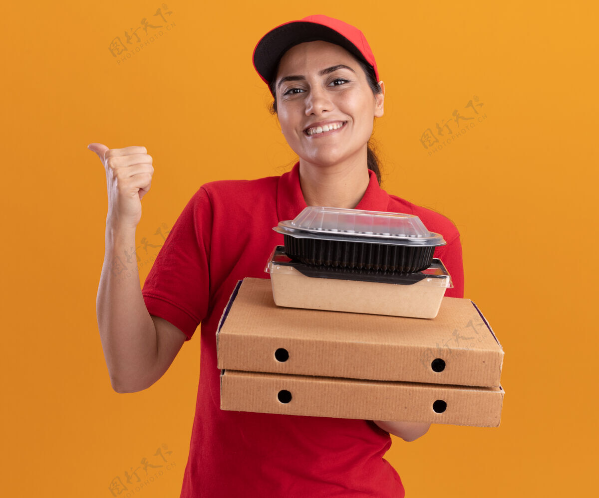 表情微笑着的年轻送货女孩穿着制服 戴着帽子 手里拿着披萨盒和食品容器 后面是橙色的隔离墙 有复印空间拿着披萨帽子
