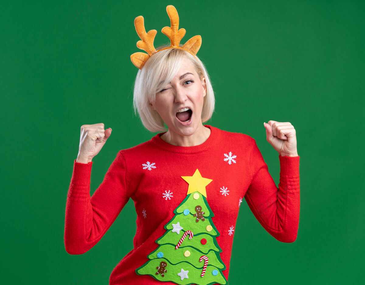 绿色快乐的中年金发女人戴着圣诞驯鹿鹿角头带和圣诞毛衣看着相机眨眼做是的手势隔离在绿色背景上手势金发中年