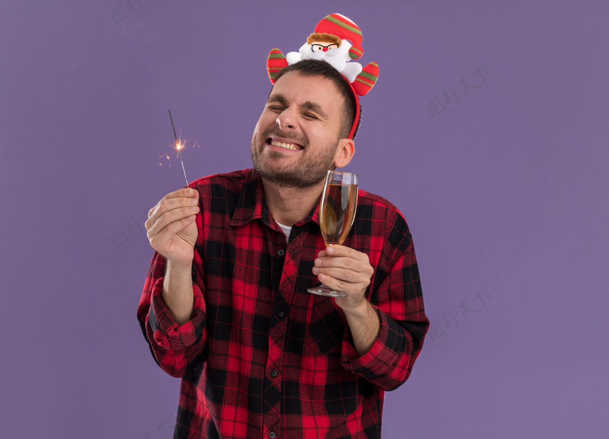 头带兴奋的白人年轻人戴着圣诞老人的头带 手里拿着节日的火花灯和香槟 闭着眼睛微笑着 紫色的背景上孤立着香槟太空紫色