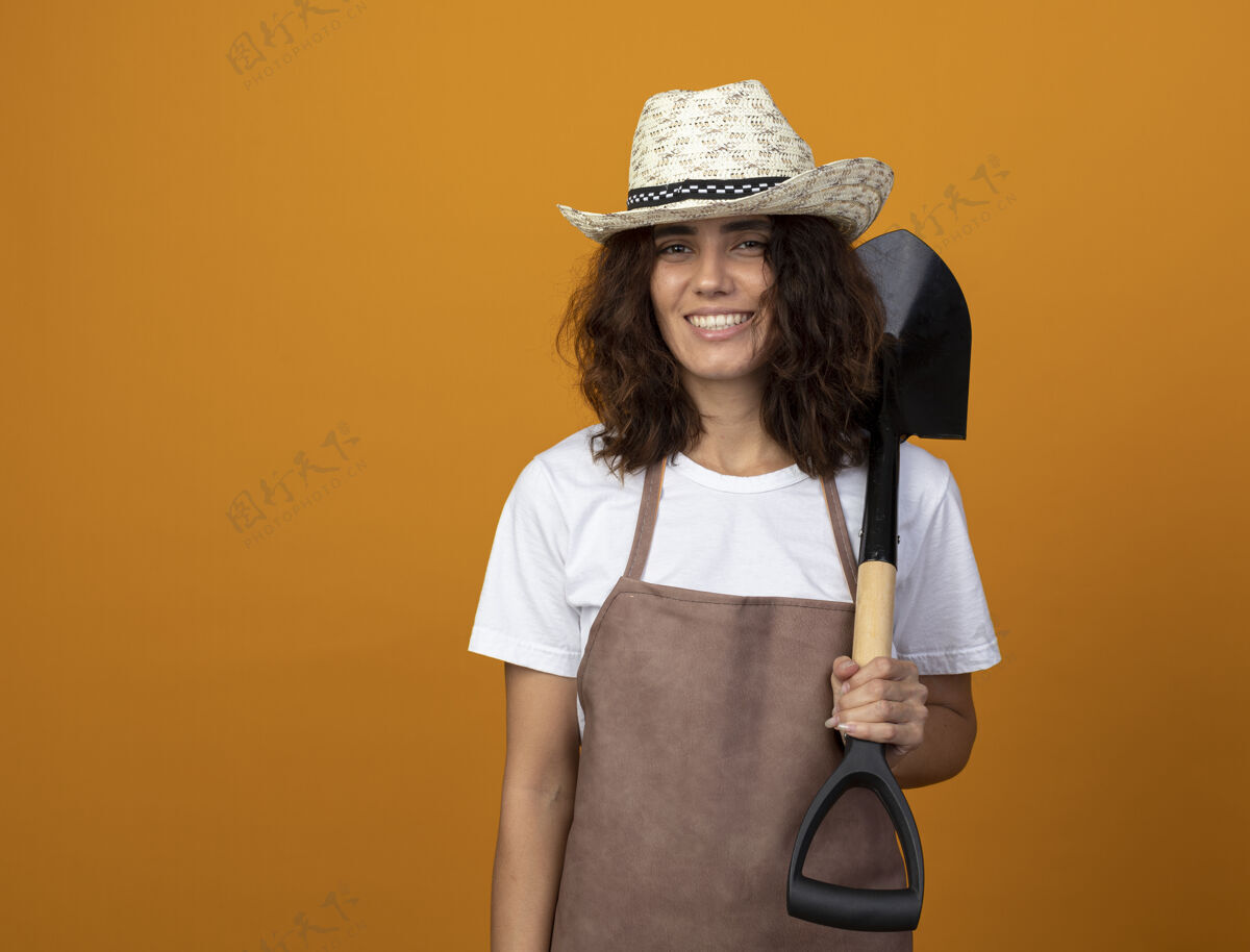 女人微笑着的年轻女园丁 穿着制服 戴着园艺帽 肩上扛着铲子举行年轻黑桃