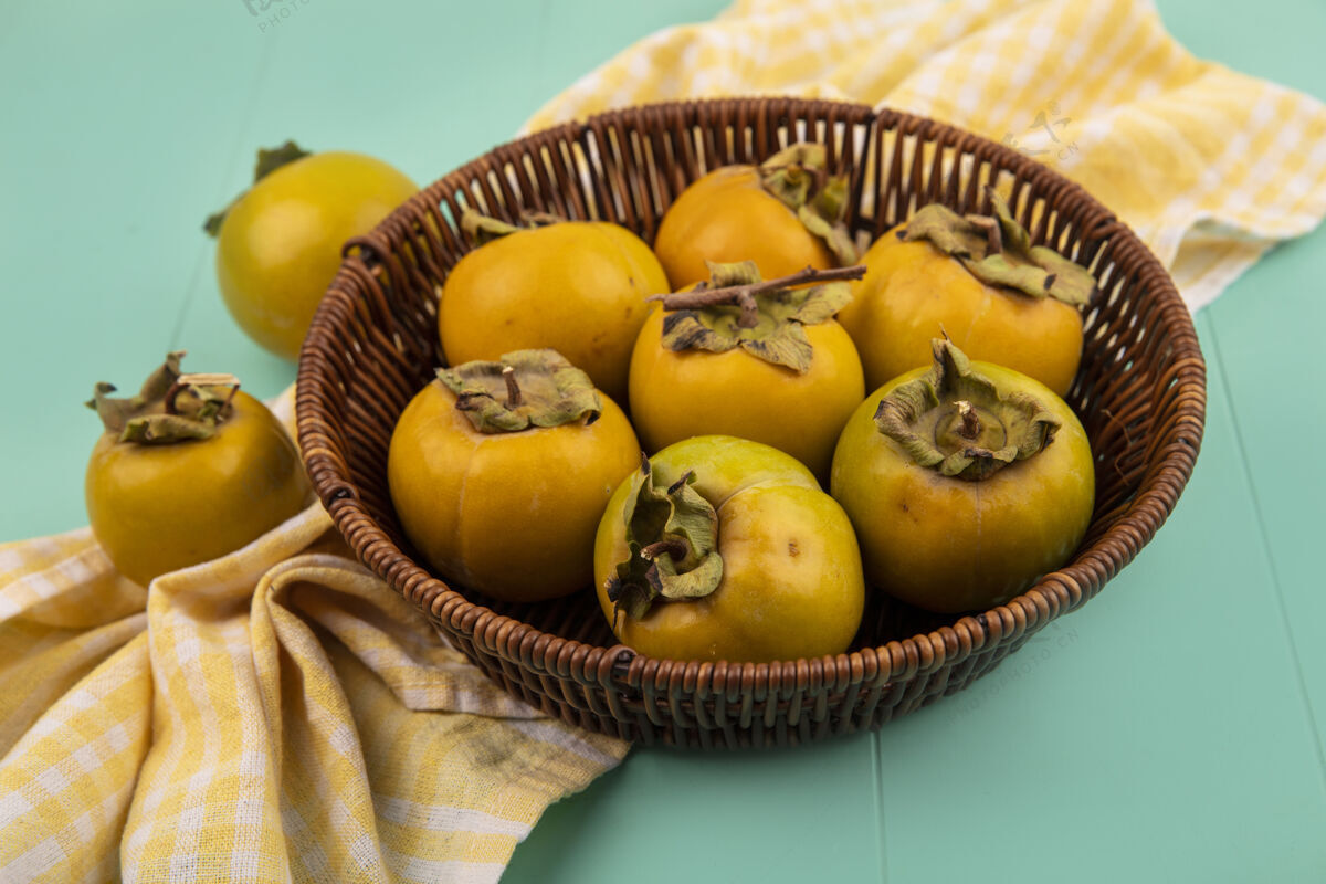 橘子蓝色木桌上黄色格子布上的水桶上新鲜未熟柿子的俯视图食物新鲜蓝色