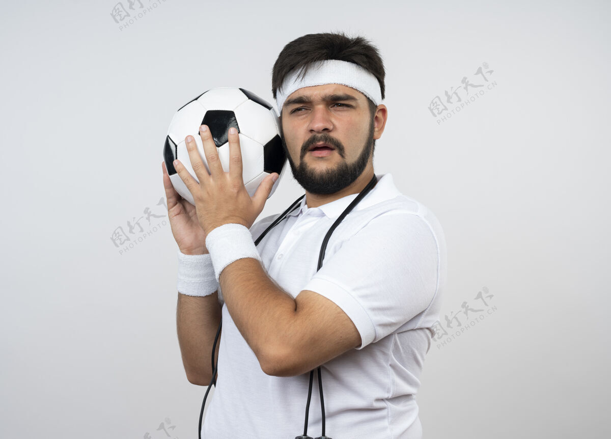 周围可疑的年轻人戴着头带和腕带 肩上带着跳绳 把球绕在隔离在白墙上的脸上抱着穿运动