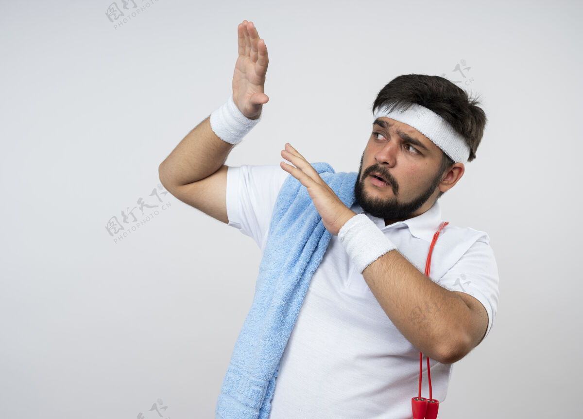 毛巾吓得年轻的运动型男子一边看着戴着头巾和手镯 一边用毛巾和跳绳搭在肩上 双手孤立地举在白色的墙上男人壁板年轻