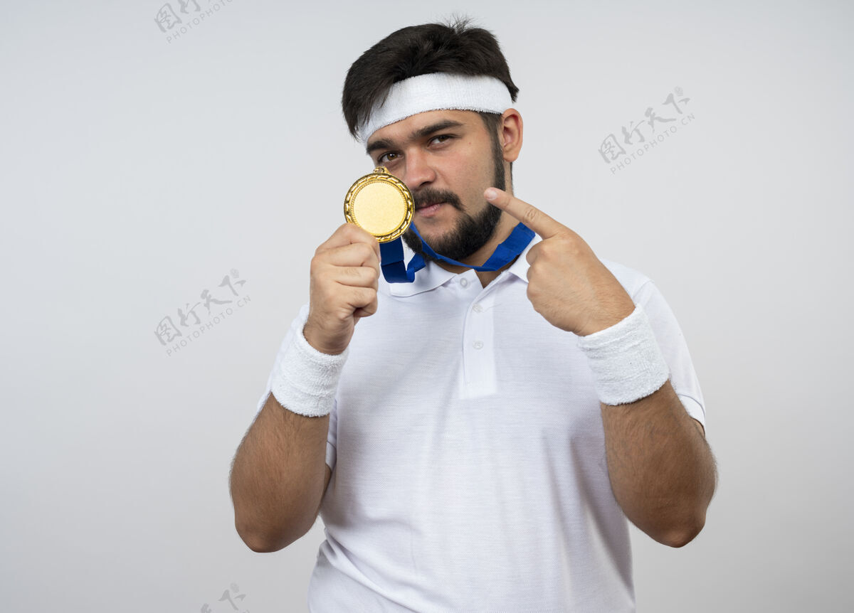 点自信的年轻人戴着头带和腕带 指着隔离在白墙上的奖牌腕带运动头带