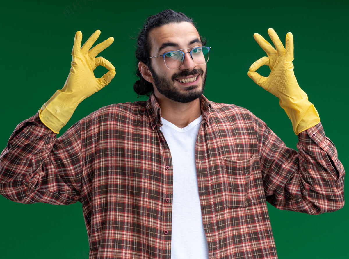 穿带着微笑的年轻帅气的清洁工 穿着t恤 戴着手套 在绿色的墙上显示出良好的姿态男人手势展示