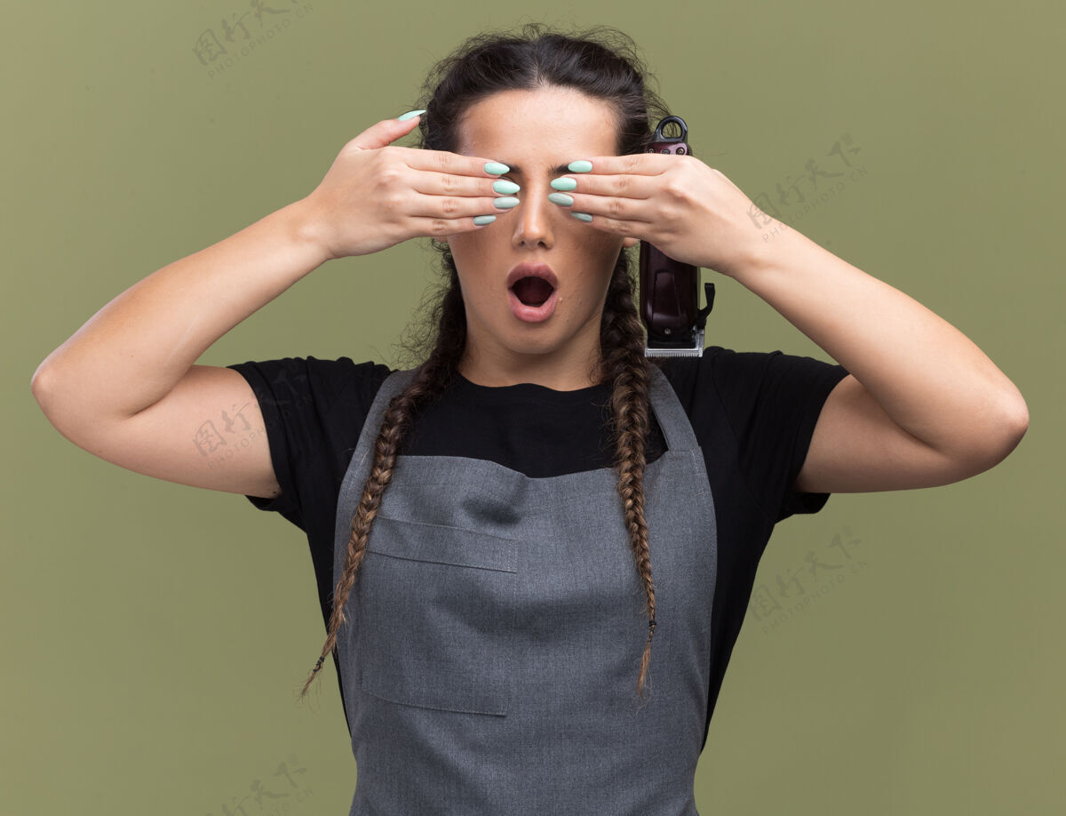 剪子惊讶的年轻女理发师穿着制服拿着理发器捂着眼睛 双手孤立在橄榄绿的墙上制服拿着橄榄