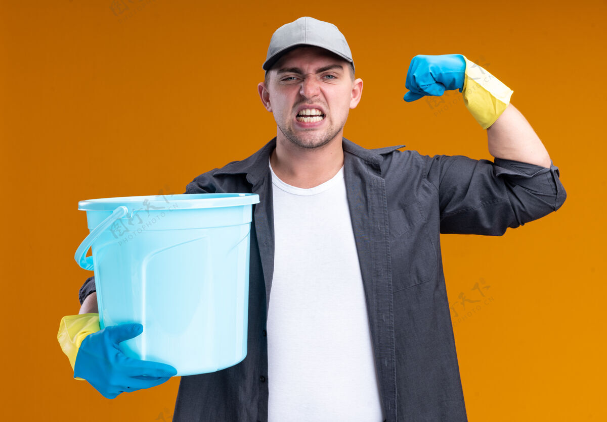 帽子自信的年轻帅气的清洁工 穿着t恤 戴着帽子 戴着手套 拿着水桶 表现出强烈的姿态 隔离在橙色的墙上抱着展示水桶