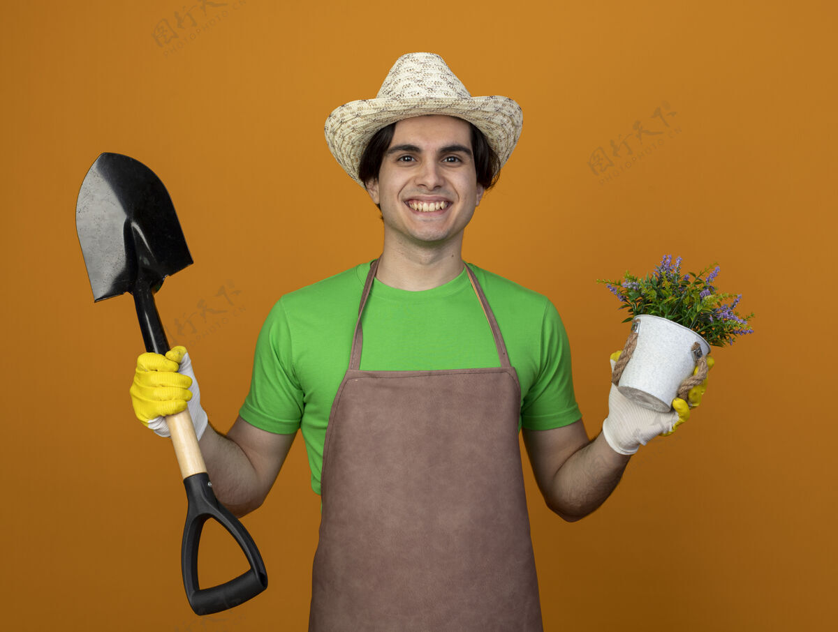 男年轻的男园丁微笑着 身着制服 戴着园艺帽 戴着手套 手里拿着铁锹 花盆里的花被隔离在橘子上花园年轻黑桃