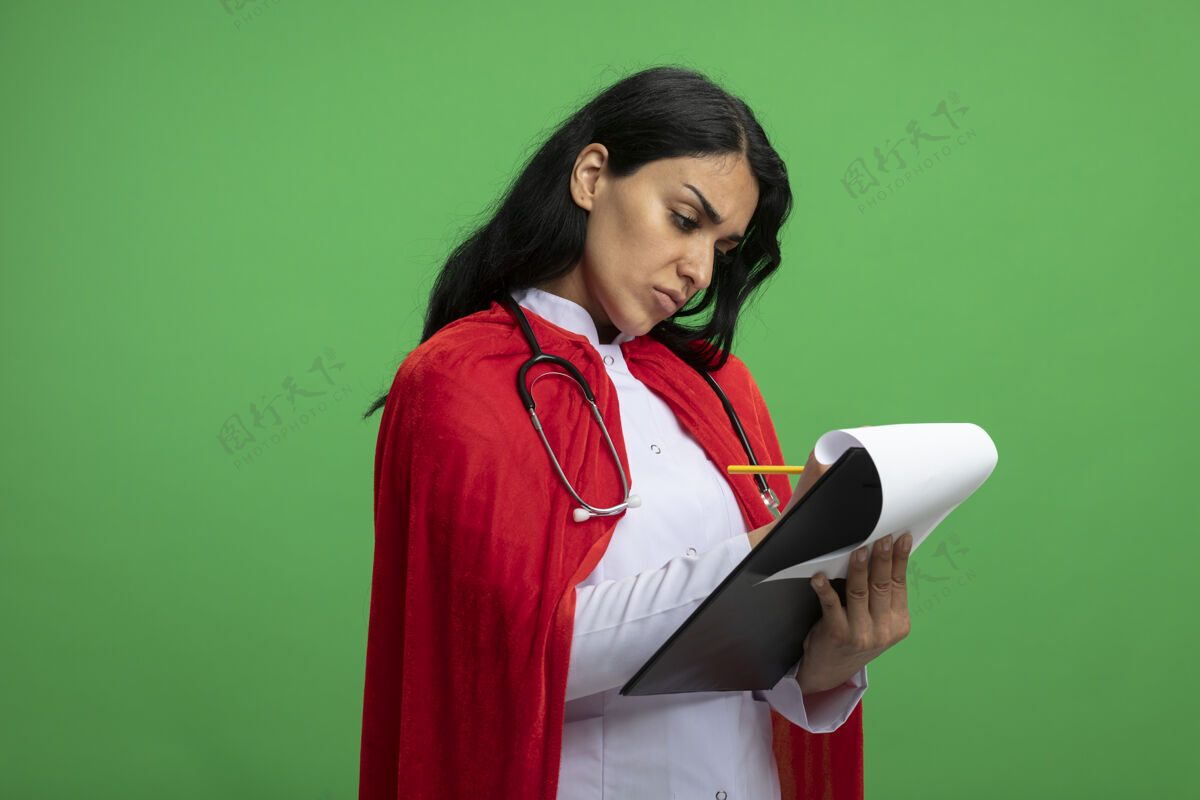 听诊器自信的年轻超级英雄女孩穿着医用长袍 手持听诊器 在绿色的剪贴板上写着什么信心写作绿色