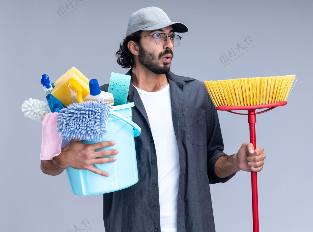 帅哥惊讶的看着身边年轻帅气的清洁工穿着t恤 戴着帽子 拿着一桶清洁工具 用拖把隔离在白色的墙上穿举行清洁