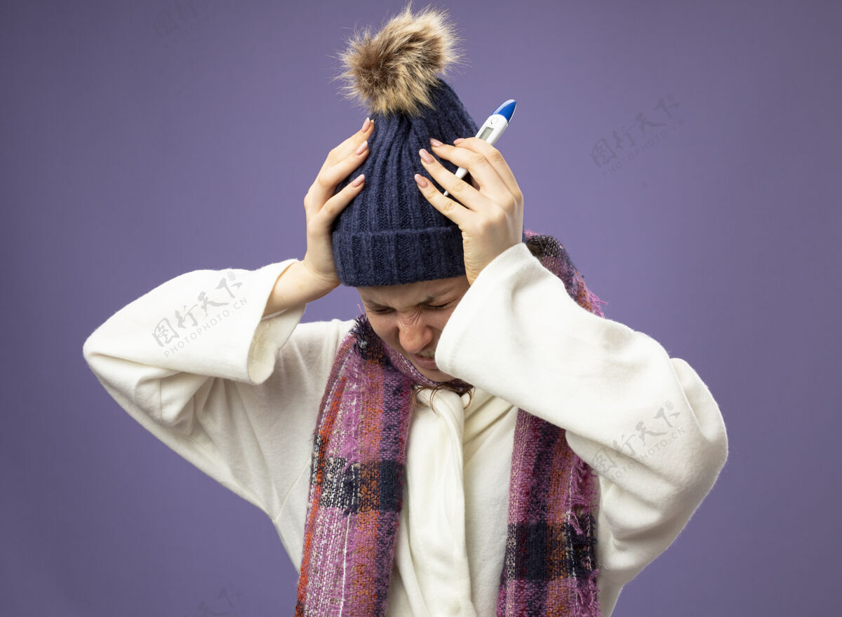 疼头痛得要命的年轻病女孩穿着白袍 戴着冬天的帽子 戴着围巾 拿着体温计 一把抓住了孤立在紫色头上的头围巾生病帽子