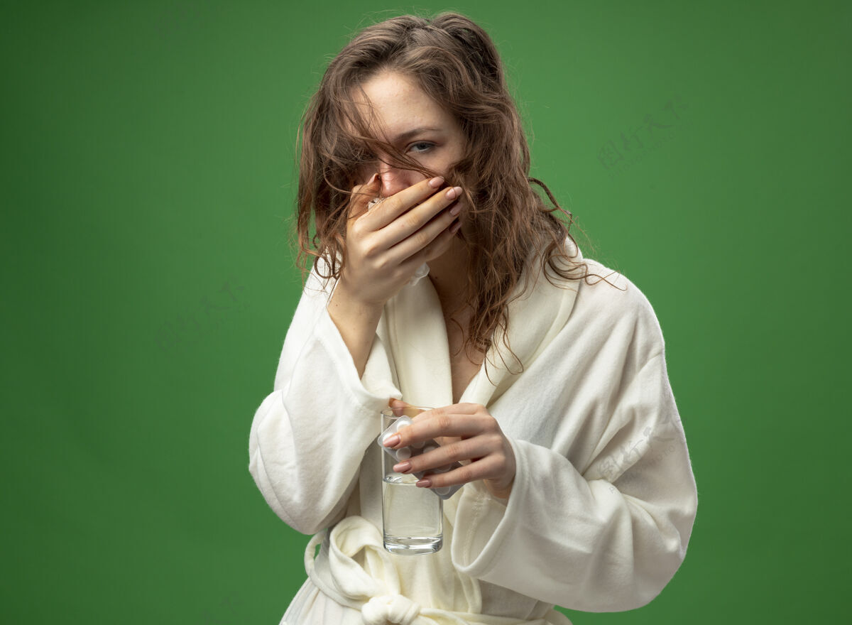 病咳嗽的年轻生病的女孩穿着白色长袍 拿着一杯水和药片 用手捂着嘴隔离在绿色上幼绿丸