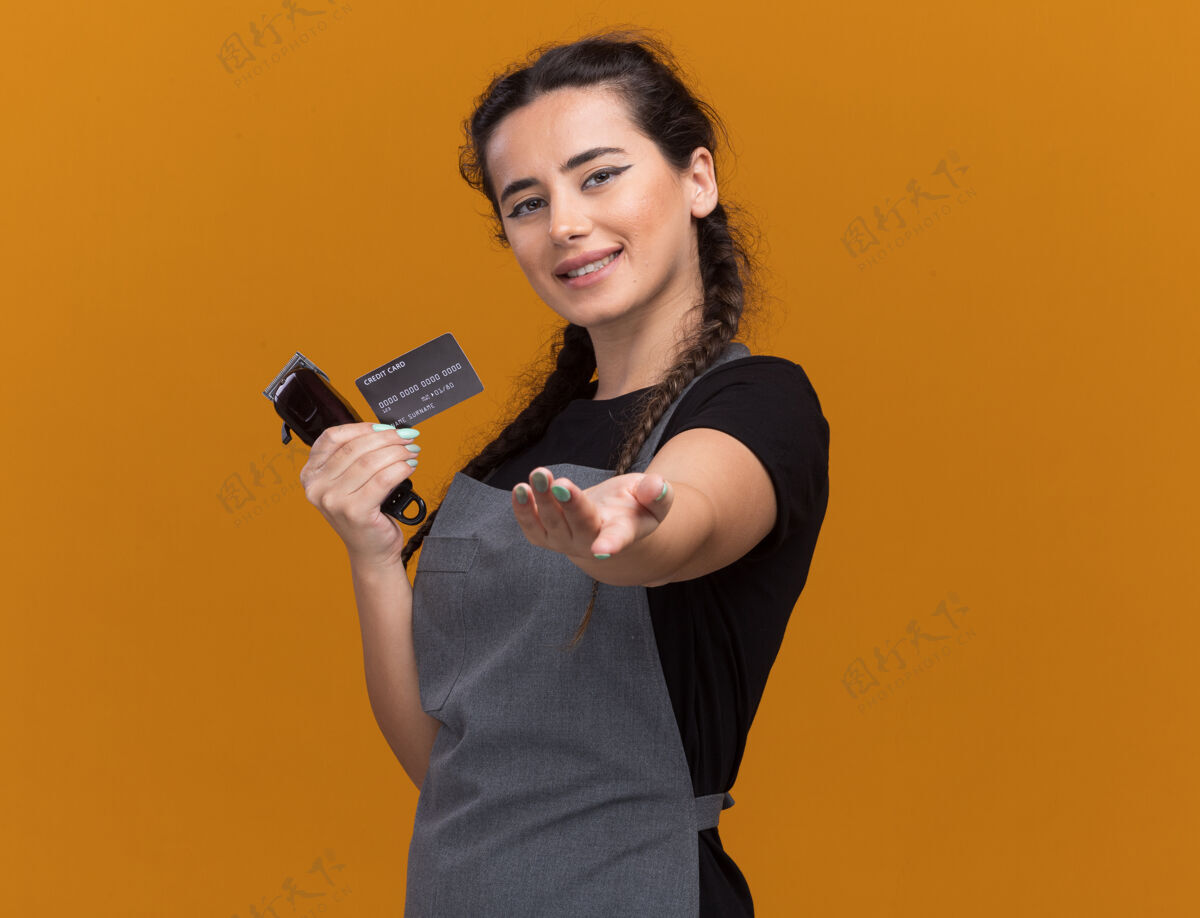 微笑微笑的年轻女理发师穿着制服 手里拿着信用卡 手里拿着理发器 手孤立地放在橙色的墙上 留着复印空间理发师信用手