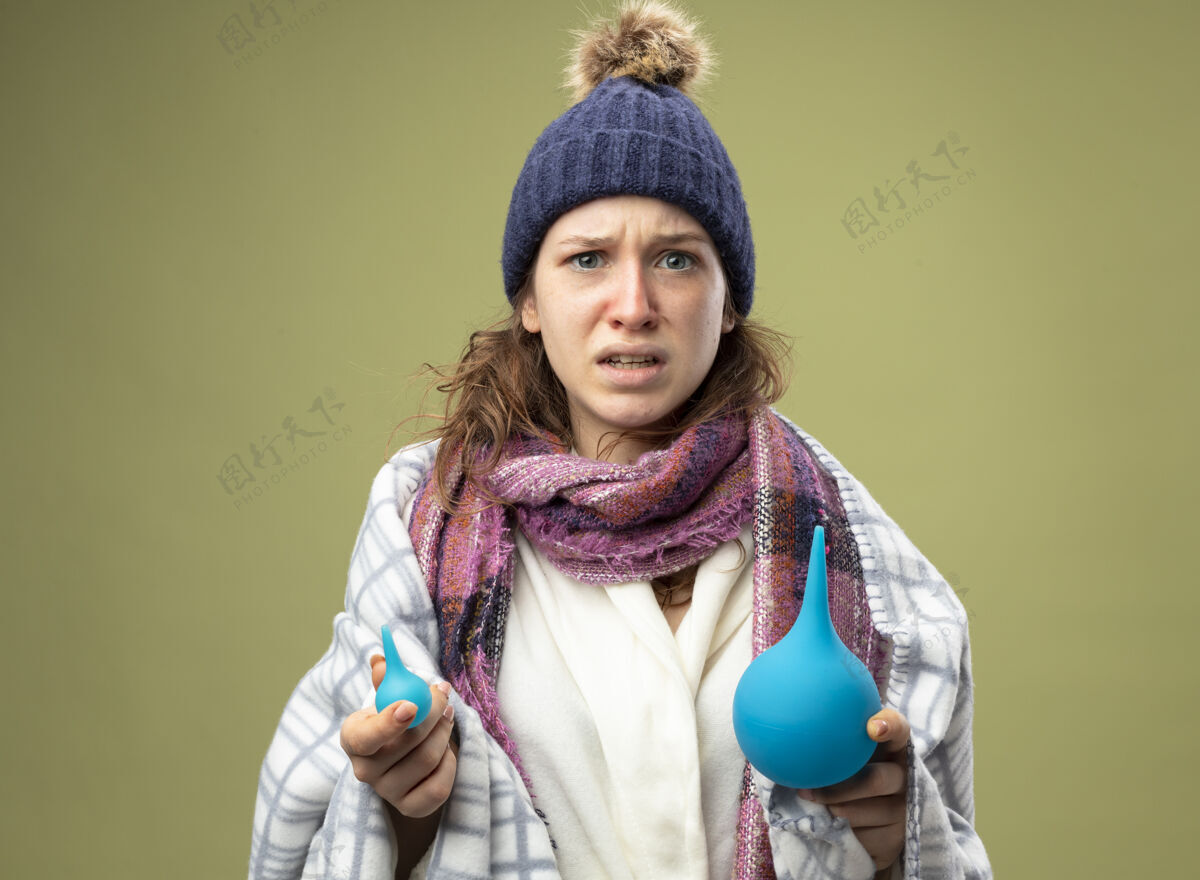 围巾困惑的年轻生病的女孩穿着白色长袍 戴着冬天的帽子 围巾裹着格子布 拿着隔离在橄榄绿上的灌肠剂包裹帽子格子