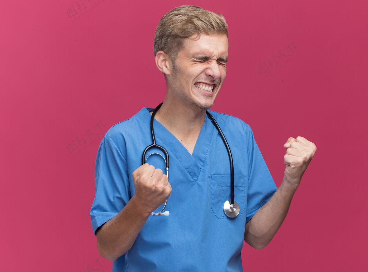 听诊器兴奋的闭着眼睛年轻男医生穿着医生制服听诊器显示是的手势隔离在粉红色的墙上手势年轻兴奋