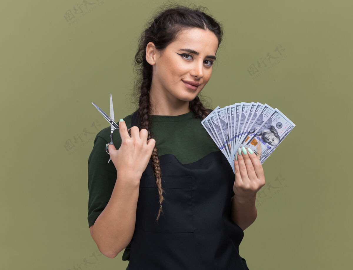 女高兴的年轻女理发师穿着制服拿着现金和剪刀隔离在橄榄绿的墙上制服理发师请拿着