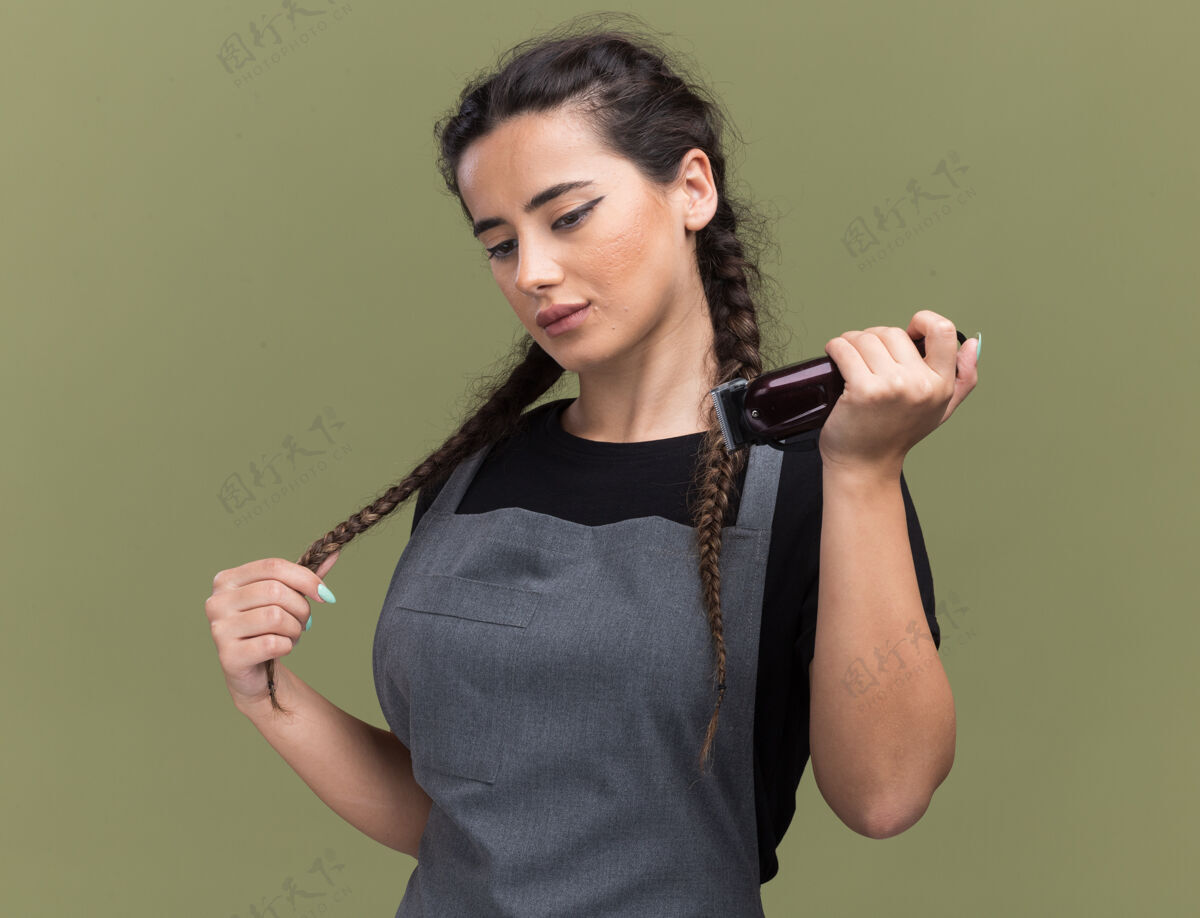 制服印象深刻的年轻女理发师穿着制服拿着理发器抓住头发孤立在橄榄绿的墙上女剪子抓取