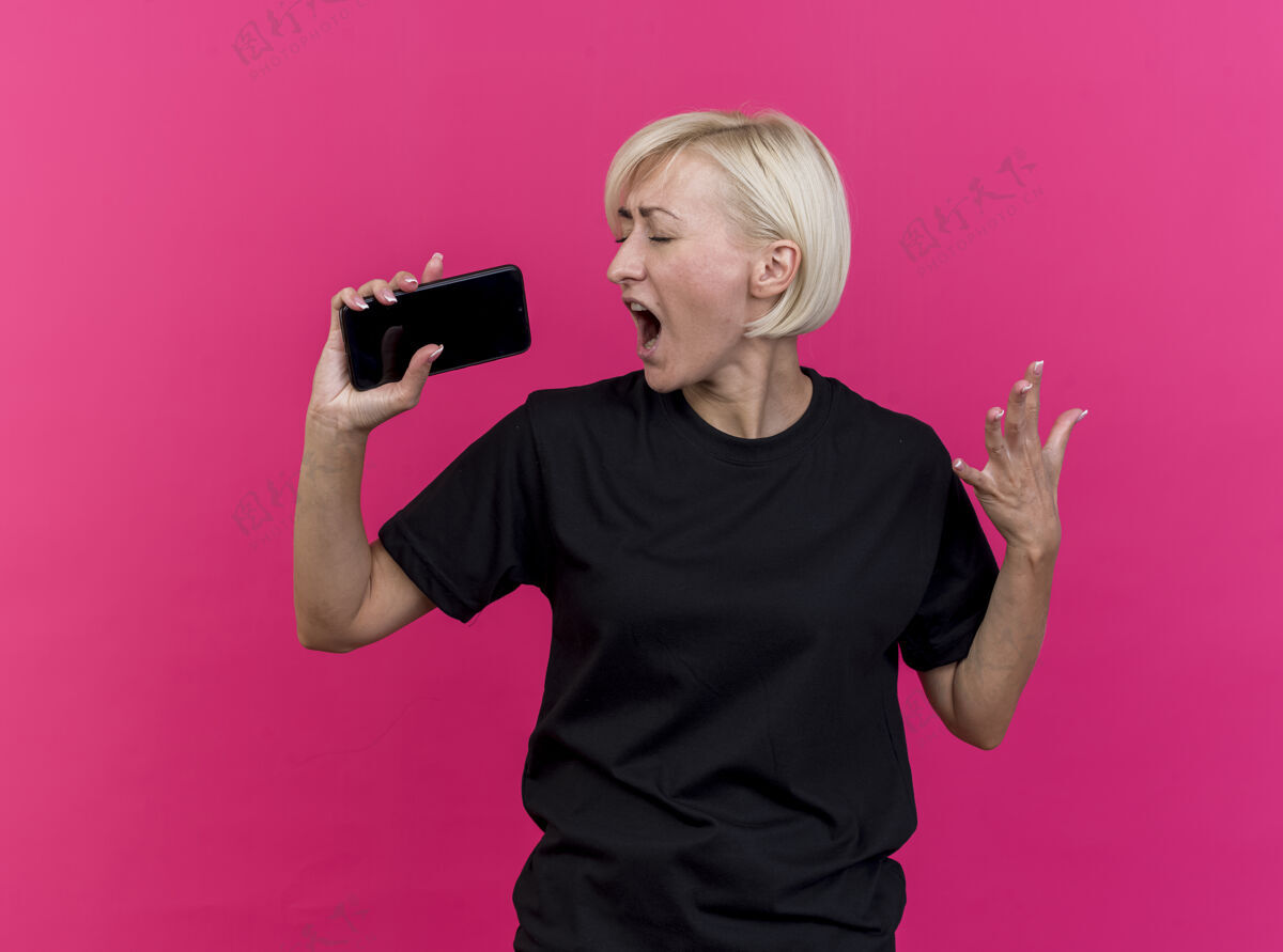 脸中年金发斯拉夫女人闭着眼睛唱歌手举在空中用手机作为麦克风隔离在粉红色的墙上市民电话手机