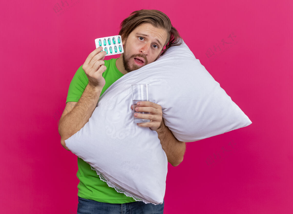 姿势印象深刻的年轻英俊的斯拉夫病人拥抱枕头看着前面举行的胶囊包和一杯水隔离在粉红色的墙上包人拥抱