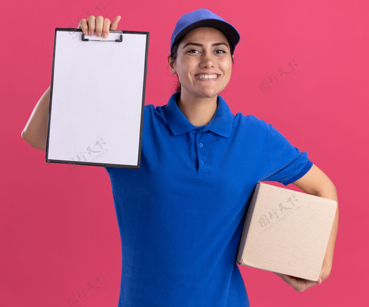 年轻带着微笑的年轻送货员 穿着制服 举着帽子 拿着一个孤立在粉红色墙上的盒子剪贴板举行制服