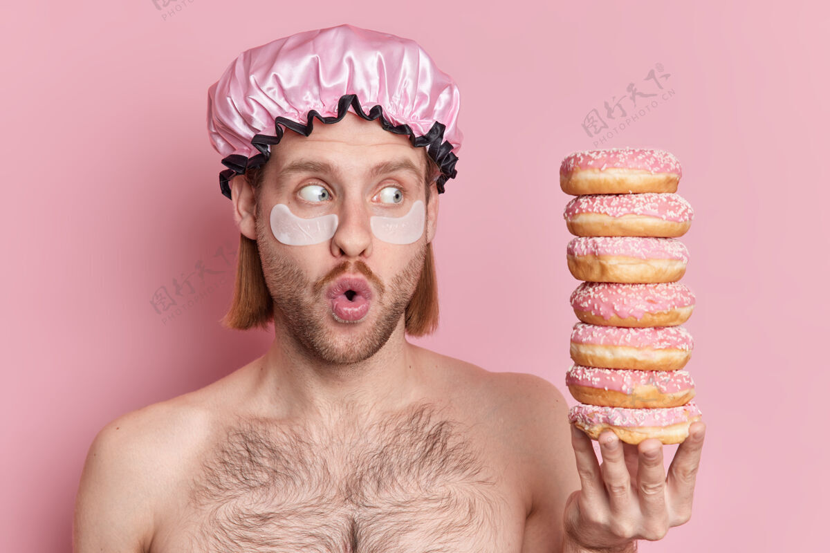 身体惊呆了的欧洲男人在眼睛下面涂上保湿垫 张着嘴盯着一堆美味的甜甜圈 光着肩膀对着粉色的工作室墙壁摆姿势男人护理卫生