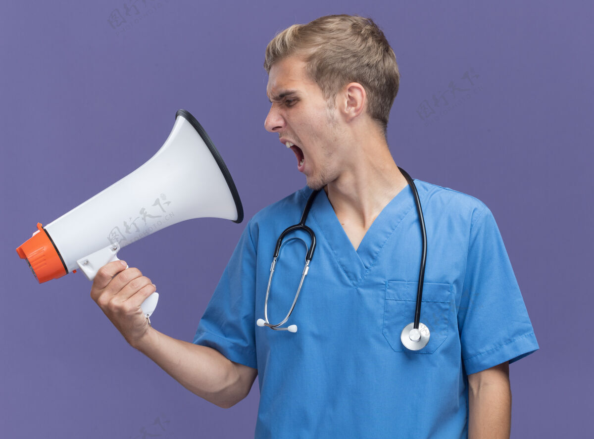 愤怒愤怒的年轻男医生穿着医生制服 戴着听诊器 用隔离在蓝色墙上的扬声器讲话听诊器医生制服