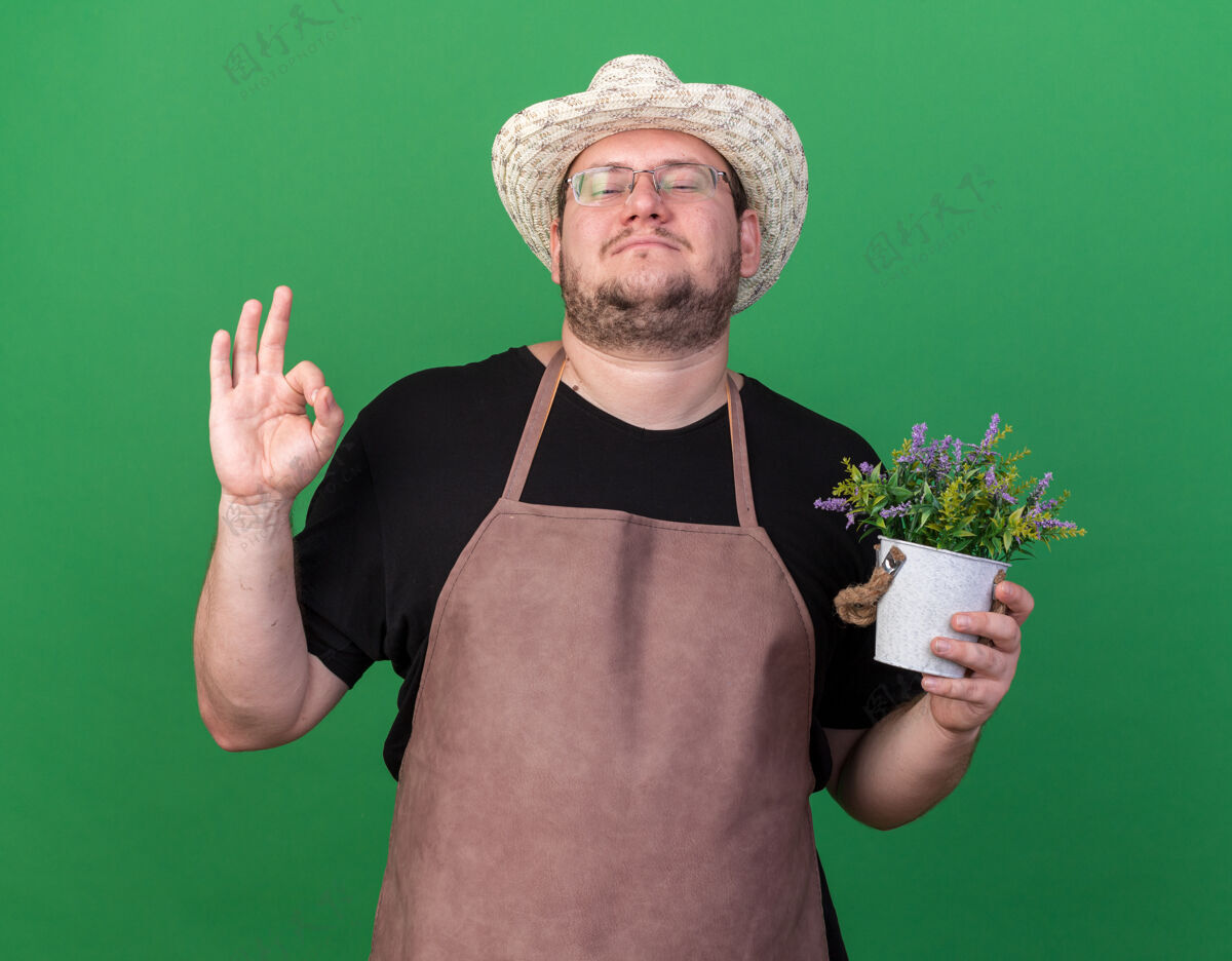 年轻自信的年轻男园丁戴着园艺帽 在花盆里捧着花 在绿色的墙上显示出良好的姿态花园男性拿着