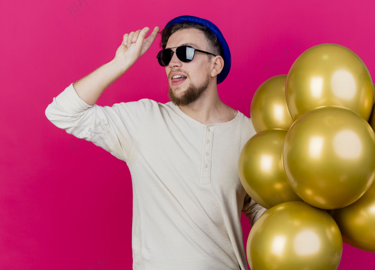 侧翼自信的年轻帅哥斯拉夫党的家伙戴着党帽和太阳镜拿着气球看着一边做失败者的手势显示舌头孤立在粉红色的墙上感觉人气球