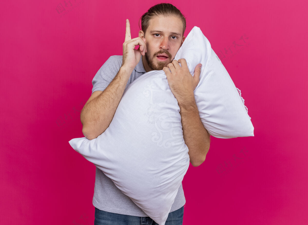 疾病印象深刻的年轻英俊的斯拉夫病夫抱着枕头看着前面举起的手指孤立地在粉红色的墙上复制空间年轻人衣服拥抱