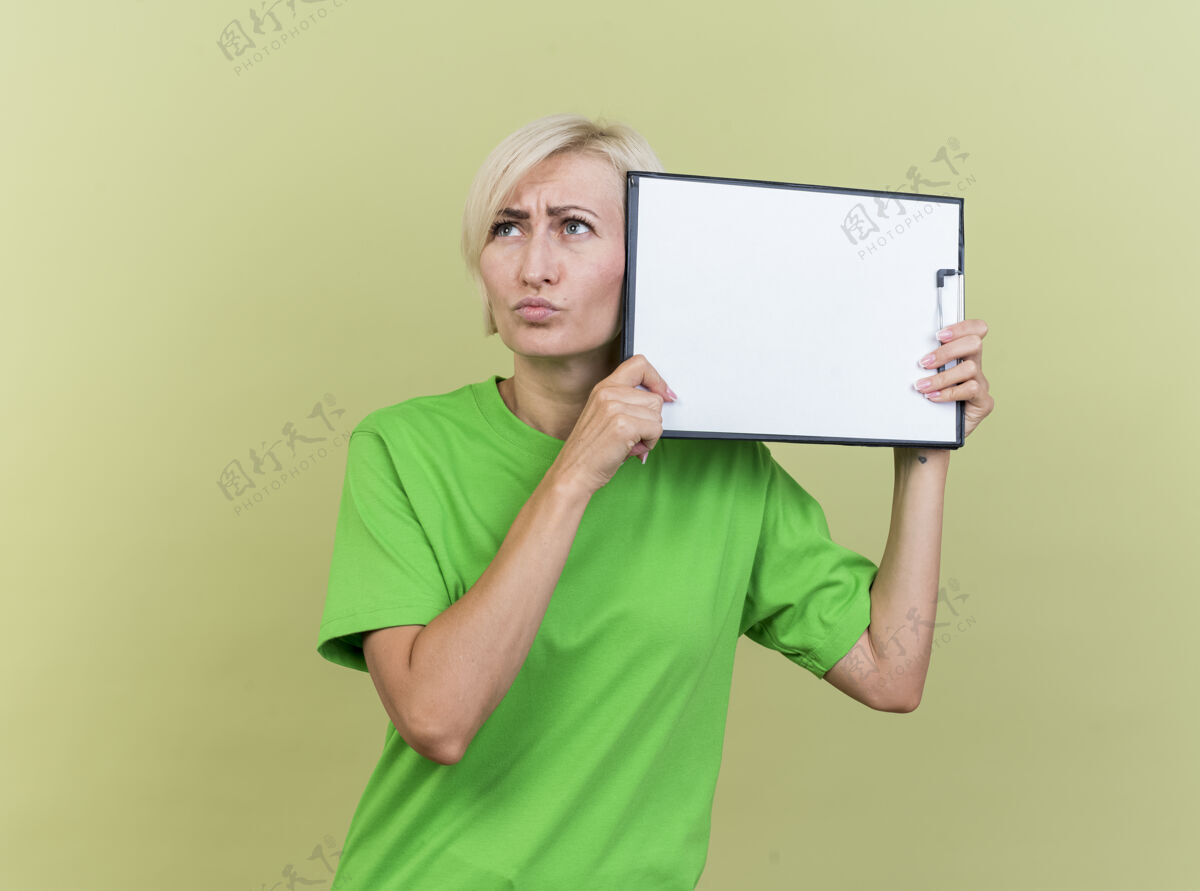 人困惑的中年金发斯拉夫妇女拿着剪贴板靠近头部 用它抚摸着头部 看着一边孤立的橄榄绿墙壁上的复制空间橄榄金发姿势