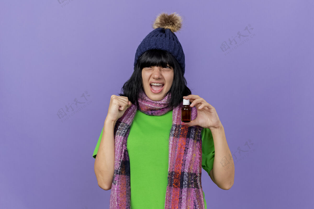 姿势戴着冬日帽 戴着围巾 手里拿着药水的快乐年轻病妇 看着一边做着“是”的手势 隔离在紫色的墙上 留着复制空间人疾病冬天