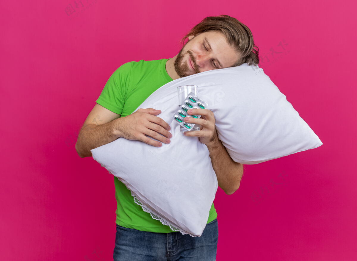 人高兴的年轻英俊的斯拉夫病夫抱着枕头把头放在上面 手里拿着一包胶囊和一杯水 看着水和胶囊隔离在粉红色的墙上 还有复制空间胶囊人头